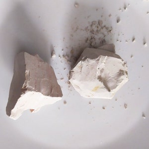 May include: Two pieces of white clay, one larger and one smaller, on a white surface. The clay is broken into irregular shapes.