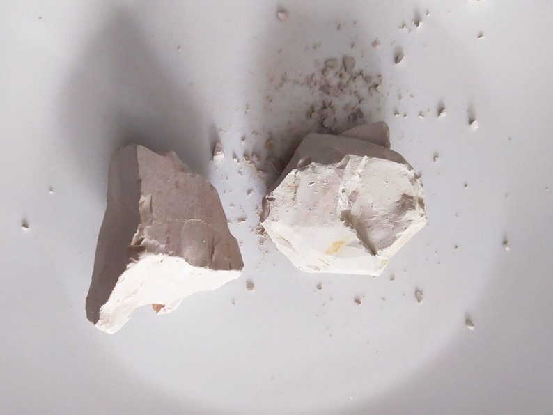 May include: Two pieces of white clay, one larger and one smaller, on a white surface. The clay is broken into irregular shapes.