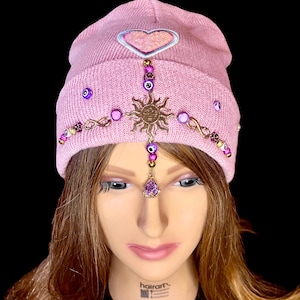 May include: A pink knit beanie with a pink heart and gold embellishments, including a sun charm, evil eye beads, and infinity symbols. The beanie is decorated with purple and pink beads.