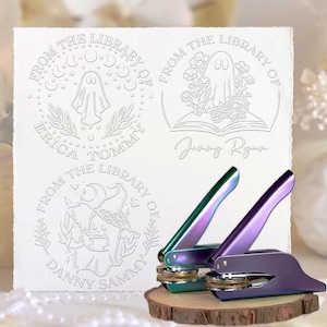 May include: Image of personalized library seals and two iridescent seal presses. The seals are embossed on white paper with ghost and witch designs. The text "FROM THE LIBRARY OF" and names are visible.