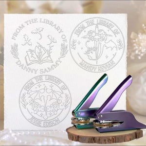 Peut inclure: Trois tampons de bibliothèque personnalisés avec des motifs détaillés et le texte "FROM THE LIBRARY OF" suivi de noms. Deux presses à cacheter irisées violettes et vertes reposent sur une base en bois, prêtes à l'emploi.