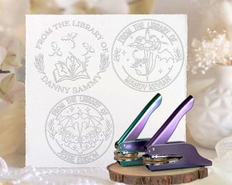 Könnte beinhalten: Drei personalisierte Bibliotheksstempel mit detaillierten Designs und dem Text "FROM THE LIBRARY OF" gefolgt von Namen. Zwei schillernde lila und grüne Siegelpressen liegen auf einem Holzsockel, gebrauchsfertig.