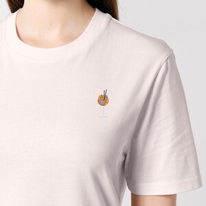 May include: A light pink t-shirt featuring a small embroidered design of an Aperol Spritz cocktail. The cocktail is in a stemmed glass with orange slices and two black straws. The shirt has a crew neck and short sleeves.
