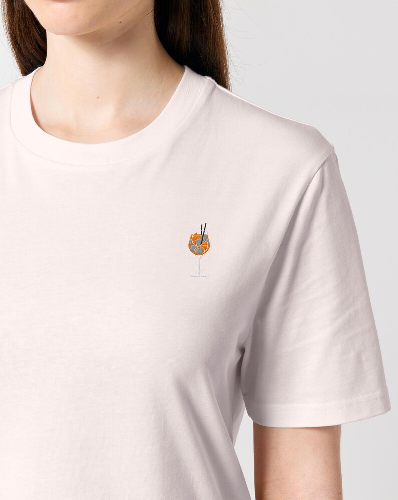 May include: A light pink t-shirt featuring a small embroidered design of an Aperol Spritz cocktail. The cocktail is in a stemmed glass with orange slices and two black straws. The shirt has a crew neck and short sleeves.