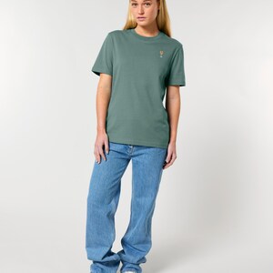 May include: A woman wearing a sage green t-shirt with a small cocktail glass graphic, paired with light blue jeans and white sneakers. The t-shirt is a solid color, and the jeans have a relaxed fit.