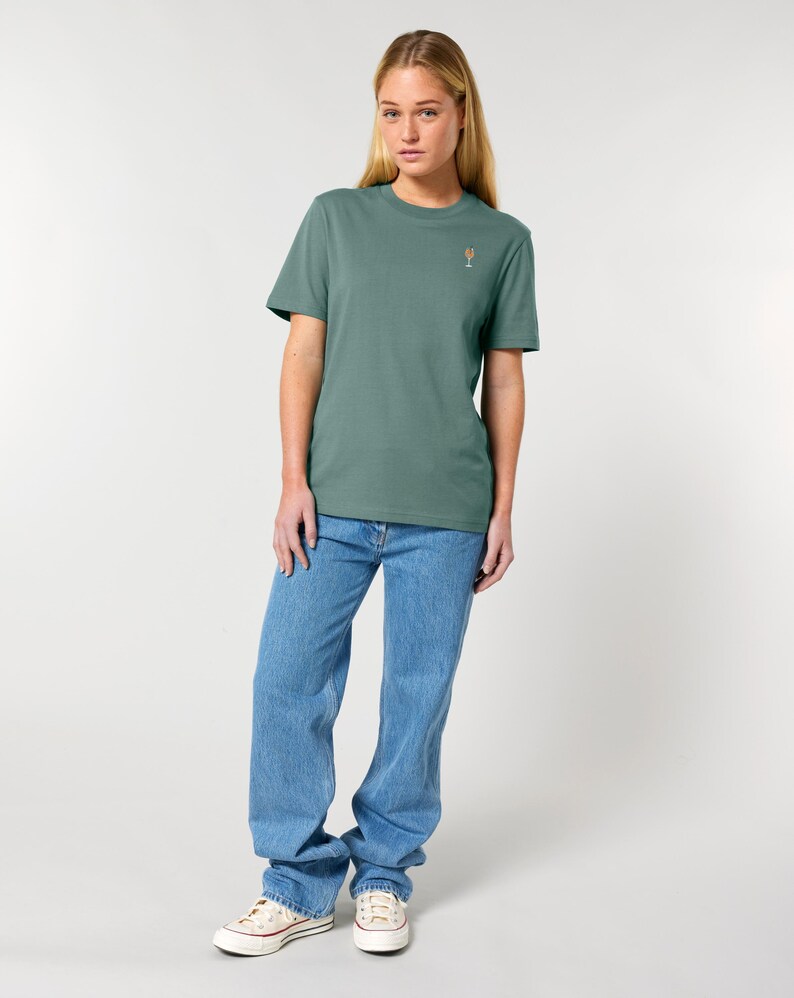 May include: A woman wearing a sage green t-shirt with a small cocktail glass graphic, paired with light blue jeans and white sneakers. The t-shirt is a solid color, and the jeans have a relaxed fit.