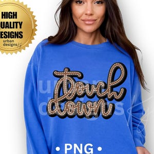 May include: A blue sweatshirt with the text "Touchdown" in a brown, stitched design. The image also includes a gold seal that says "High Quality Designs". Below the text is the word "PNG".