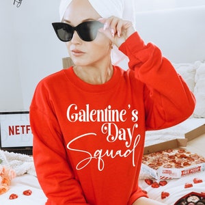 Puede incluir: Una sudadera roja con el texto "Galentine's Day Squad" en blanco. La sudadera la lleva puesta una persona con una toalla blanca en la cabeza y gafas de sol negras.