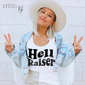 May include: A white vest top with the words "Hell Raiser" in black, worn with a light blue denim jacket with fringe. The image also shows a white hat and a white brick wall background.