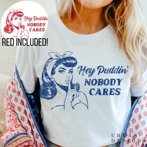 May include: White t-shirt with a blue graphic of a woman with a finger to her lips, and the text "Hey Puddin' Nobody Cares." A red graphic with the text "Hey Puddin' Nobody Cares" is in the upper left corner. The words "RED INCLUDED!" are below the graphic.