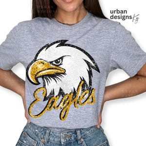 May include: A gray t-shirt with a glittery white and black eagle head design and the word "Eagles" in gold glitter lettering.