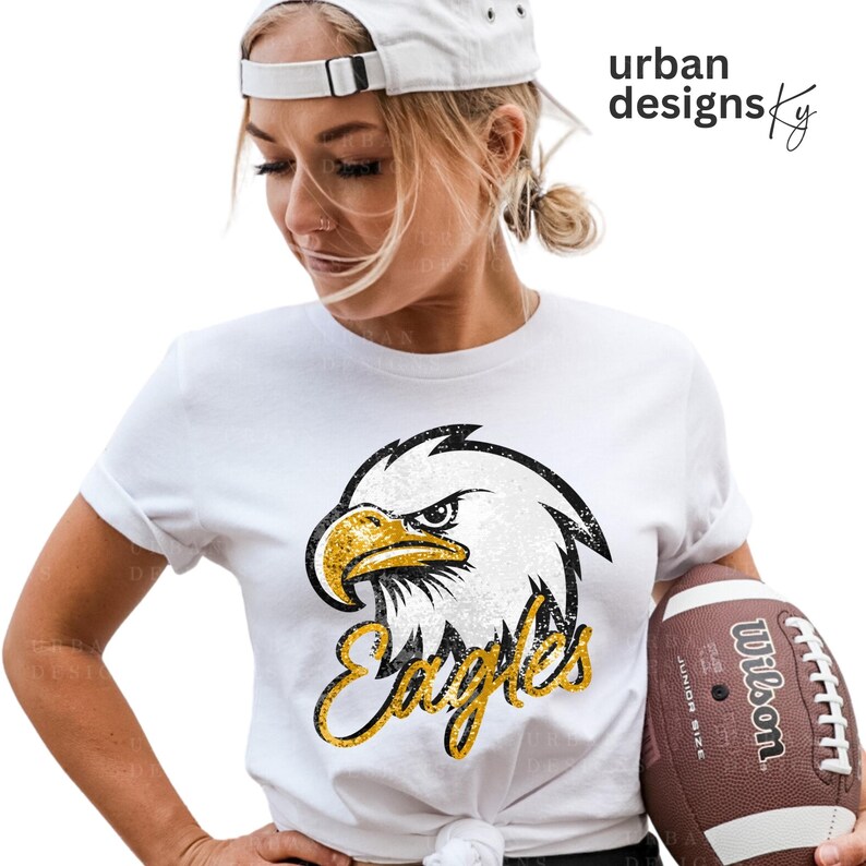 May include: White t-shirt with a black and white eagle graphic and gold glitter text that says "Eagles".