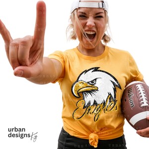 May include: A woman wearing a yellow t-shirt with a white and gold glitter eagle graphic and the word "Eagles" in gold glitter. She is holding a brown and white football with the word "Wilson" on it.
