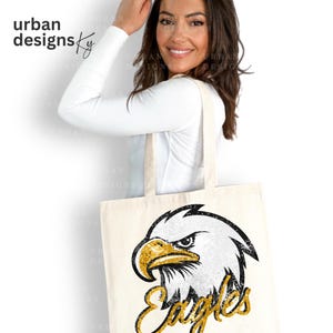 May include: A white canvas tote bag with a black and white eagle graphic and gold glitter accents. The text "Eagles" is written in gold glitter below the eagle.