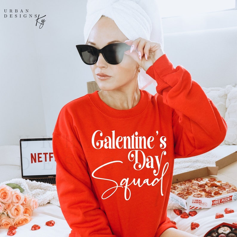 May include: A red sweatshirt with the text "Galentine's Day Squad" in white. The sweatshirt is worn by a person with a white towel on their head and black sunglasses.