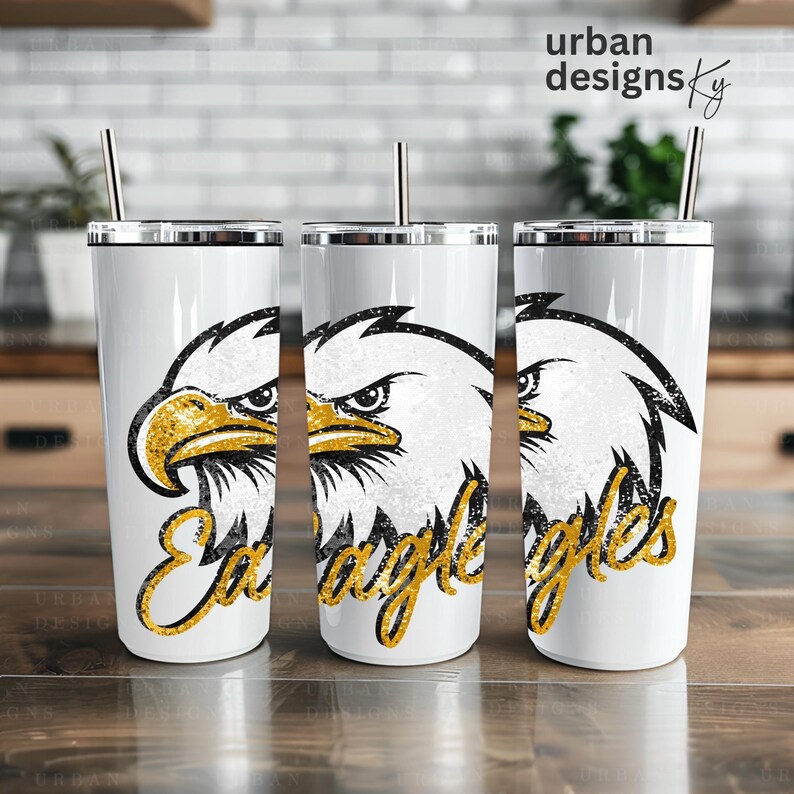 May include: Three white tumblers with a gold glitter eagle design and the word "Eagles" in gold glitter. The tumblers have a silver lid and a straw.