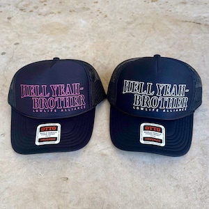 Pode incluir: Dois bonés trucker pretos com parte traseira em rede e frente em espuma. O boné da esquerda tem o texto rosa HELL YEAH-BROTHER LOWLIFE ALLIANCE, o da direita tem o mesmo texto em branco. Ambos têm um autocolante da marca OTTO na pala.