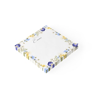 以下が含まれることがあります： 青と黄色の花柄の縁取りが施された白いメモ帳。メモ帳の前面には「A note from Emma」の文字が書かれている。