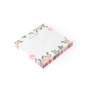 Puede incluir: Bloc de notas cuadrado con un diseño floral. El bloc de notas presenta un fondo blanco con un borde de flores y hojas rosas y verdes en acuarela. El texto "A note from Amanda" está impreso en escritura negra.