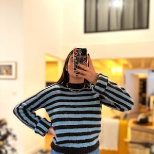 Peut inclure: Un pull en crochet à rayures bleues et marron avec des manches longues. Le pull a un col montant et est associé à un jean bleu. Un collier en or est visible.
