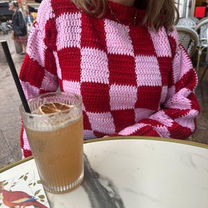 Peut inclure: Un pull crocheté à motif damier rouge et rose. Un grand verre d'une boisson brun clair avec une paille et une tranche d'orange séchée en garniture est posé sur une table en marbre.