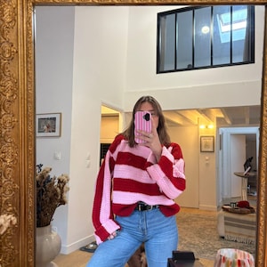 Könnte beinhalten: Ein rot-rosa gestreifter Häkelpullover mit langen Ärmeln. Der Pullover wird mit einer blauen Jeans und einem schwarzen Gürtel kombiniert. Das Bild spiegelt sich in einem goldgerahmten Spiegel.