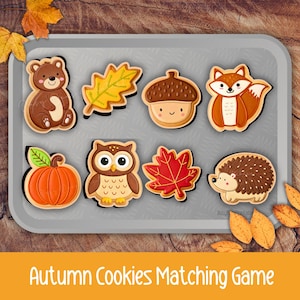 May include: A set of eight autumn-themed cookie-shaped cutouts arranged on a gray tray, featuring a brown bear, yellow oak leaf, brown acorn, orange fox, orange pumpkin, brown owl, red maple leaf, and a brown hedgehog with the text Autumn Cookies Matching Game.