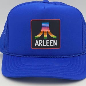 May include: A royal blue trucker hat with a mesh back and a curved brim. The front features a rectangular patch with a colorful retro design and the name "ARLEEN" in white letters.