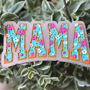 May include: A decorative sign with the word "MAMA" in large, stylized letters. The letters are turquoise with a floral pattern in pink and white, outlined in orange. The sign is set against a backdrop of green foliage.