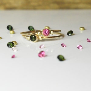 Op de afbeelding: Twee gouden ringen met een roze en groene edelsteen in elke ring. De ringen zijn omringd door losse roze en groene edelstenen.