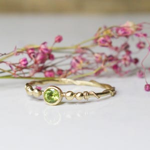 Può includere: Un delicato anello d'oro con una fascia attorcigliata e una singola pietra preziosa verde. L'anello è adornato con piccole perle dorate.