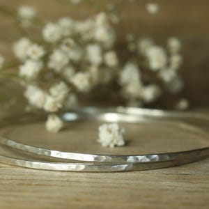 Pode incluir: Duas pulseiras de prata martelada são exibidas em uma superfície de madeira, com um fundo desfocado de flores brancas. As pulseiras têm uma superfície texturizada e um design simples e elegante, perfeito para o uso diário.