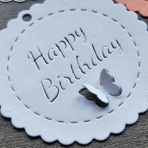May include: White round paper tag with scalloped edges and a white string attached. The tag has a dashed line border and the words "Happy Birthday" written in a cursive font. Two white paper butterflies are attached to the tag.