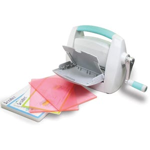 以下が含まれることがあります： 白とターコイズブルーの紙用手動トリマー。グレーのトレイとクランクハンドルが付いています。トリマーは白い表面に置かれており、ピンク、オレンジ、イエローのプラスチックシートが3枚重ねられています。