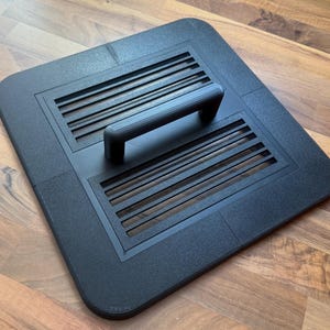 Könnte beinhalten: Eine quadratische schwarze Abdeckung mit abgerundeten Ecken, einem mittigen Griff und zwei rechteckigen Lüftungsbereichen mit horizontalen Lamellen. Die Oberfläche ist texturiert und das Design weist eine deutliche Naht in der Mitte auf.