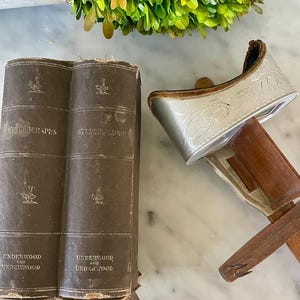 May include: An antique stereoscope with a silver-colored viewer and wooden handle, next to two brown books titled "Stereographs" and "Underwood and Underwood". The books and viewer are placed on a white marble surface.