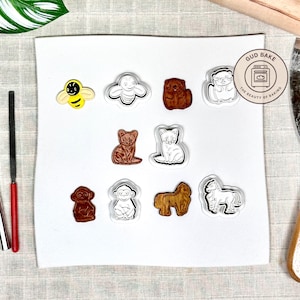 Könnte beinhalten: Eine Sammlung von Ausstechern in Tierform, darunter eine Biene, ein Schmetterling, ein Bär, eine Katze, ein Affe, ein Hund und ein Pferd. Die Ausstecher sind in verschiedenen Farben und Designs gehalten, mit weißem Umriss. Das Bild enthält den Text "Gud Bake".