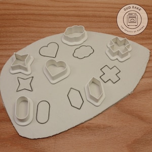 Könnte beinhalten: Ein Set mit 10 weißen Plastik-Ausstechern auf einer grauen Oberfläche. Die Ausstecher sind in Form eines Sterns, Herzens, einer Wolke, eines Kreuzes, eines Ovals und eines Sechsecks. Die Ausstecher sind in einem Muster auf der Oberfläche angeordnet. Der Text "GUD BAKE THE BEAUTY OF BAKING" ist in einem braunen Kreis in der oberen rechten Ecke des Bildes sichtbar.