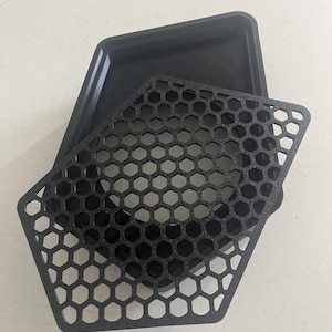 Puede incluir: Una bandeja pentagonal negra con una superficie interior lisa y un inserto pentagonal negro a juego que presenta un patrón de rejilla de panal texturizado. El inserto descansa parcialmente dentro de la bandeja, mostrando el diseño geométrico.