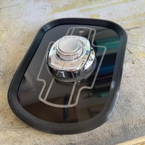 May include: A black, rectangular car console control panel with a chrome-finished control knob and buttons. The panel has a raised black border and a silver-colored design. The control knob is in the center, surrounded by buttons with symbols.
