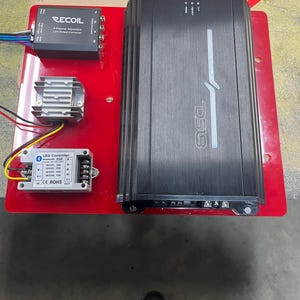 May include: A black car amplifier with the text "D518" on the front. The amplifier is mounted on a red surface with a black LED controller and a black line output converter. The text on the LED controller reads "LED Controller Bluetooth 12V DC 5A".