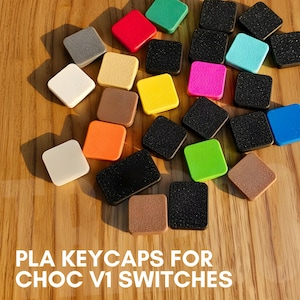 May include: A selection of square keycaps in a variety of colours, such as red, green, yellow, and black, arranged on a wooden surface. The text at the bottom reads "PLA KEYCAPS FOR CHOC V1 SWITCHES."