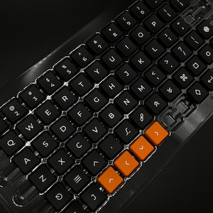 Pode incluir: Um conjunto de teclas de teclado pretas com letras brancas e teclas de seta laranja, dispostas numa bandeja de plástico transparente. Disposição QWERTY. A marca "TRAPDOG" é visível.