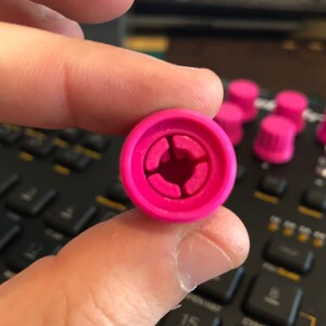 Peut inclure: Gros plan sur un bouton de commande rose vif, avec un design circulaire et une ouverture centrale. Le bouton est tenu entre les doigts, avec d'autres boutons similaires visibles en arri&egrave;re-plan, sugg&eacute;rant un ensemble de composants d'&eacute;quipement audio.