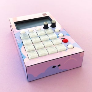 以下が含まれることがあります： ピンクとパープルの音楽サンプラー。白いボタンのグリッドと2つのグレーのノブがあります。サンプラーは黒い縁取りと上部に画面があります。