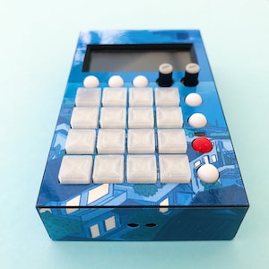 May include: A blue and white electronic music sampler with a grid of 16 buttons and a small screen. The sampler has a cityscape design with buildings and trees.