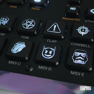 以下が含まれることがあります： 黒色の電子音楽制作デバイスのクローズアップ。ボタンは、五芒星、悪魔の顔、ストームトルーパーのヘルメットなどのアイコンを表示します。「Clear」、「Paste」、「Tom」、「Clap」、「Cowbell」などのテキストが含まれています。「TRAPDOG」のブランド名が表示されています。