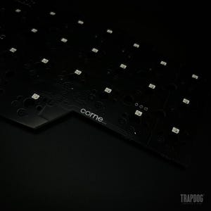 May include: Close-up of a black circuit board with numerous small, square LED lights. The board has a complex pattern of circuits and components, with the word "corne" printed on it. The overall aesthetic is dark and technical.