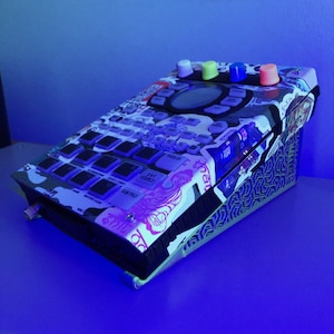 以下が含まれることがあります： カラフルなステッカーデザインが施された白黒のドラムマシン。ドラムマシンには、ホールドボタン、エグジットソースボタン、サーパッドボタンなど、さまざまなボタンとノブがあります。ドラムマシンは青い表面の上に置かれています。