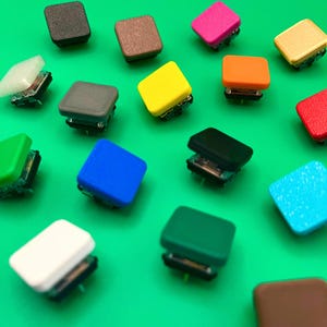 Può includere: Un assortimento di pulsanti quadrati colorati in varie tonalità, tra cui nero, marrone, rosa, giallo, arancione, rosso, verde, blu, bianco e azzurro. I pulsanti sono disposti su una superficie verde brillante, mostrando i loro diversi colori e design.